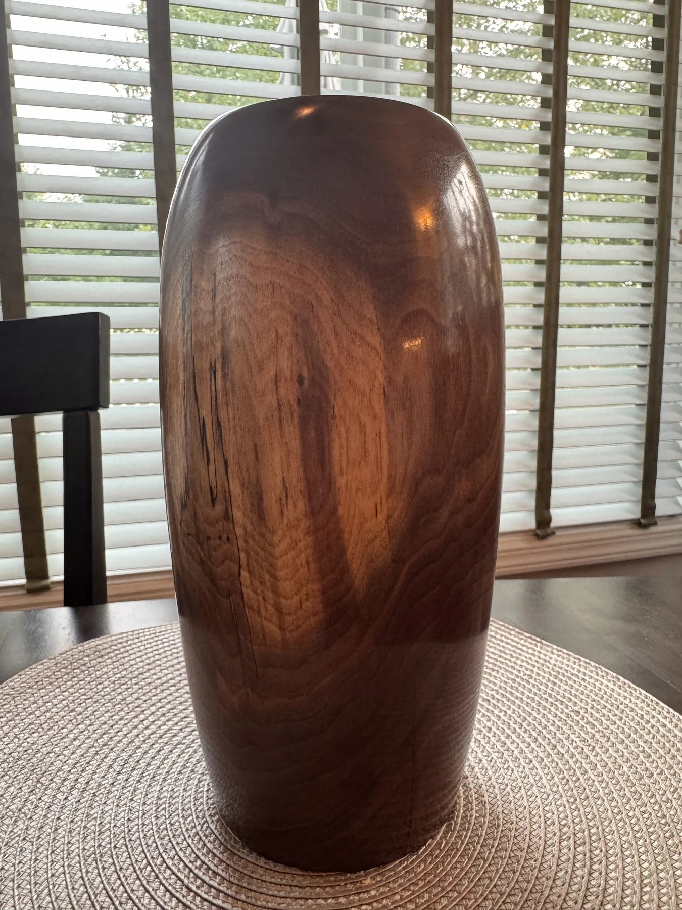 Walnut vase on a woven dining-table mat with window blinds behind it